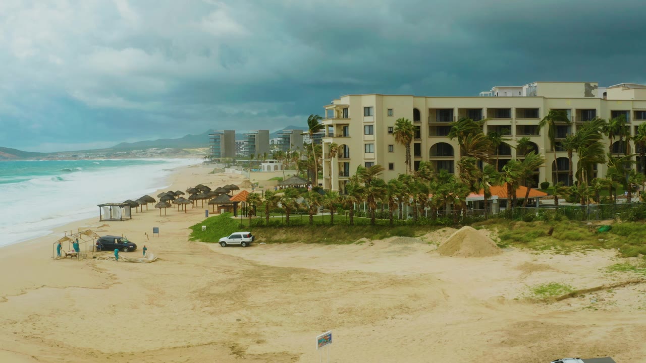 Sandy beach with resort building, cars, and waves.