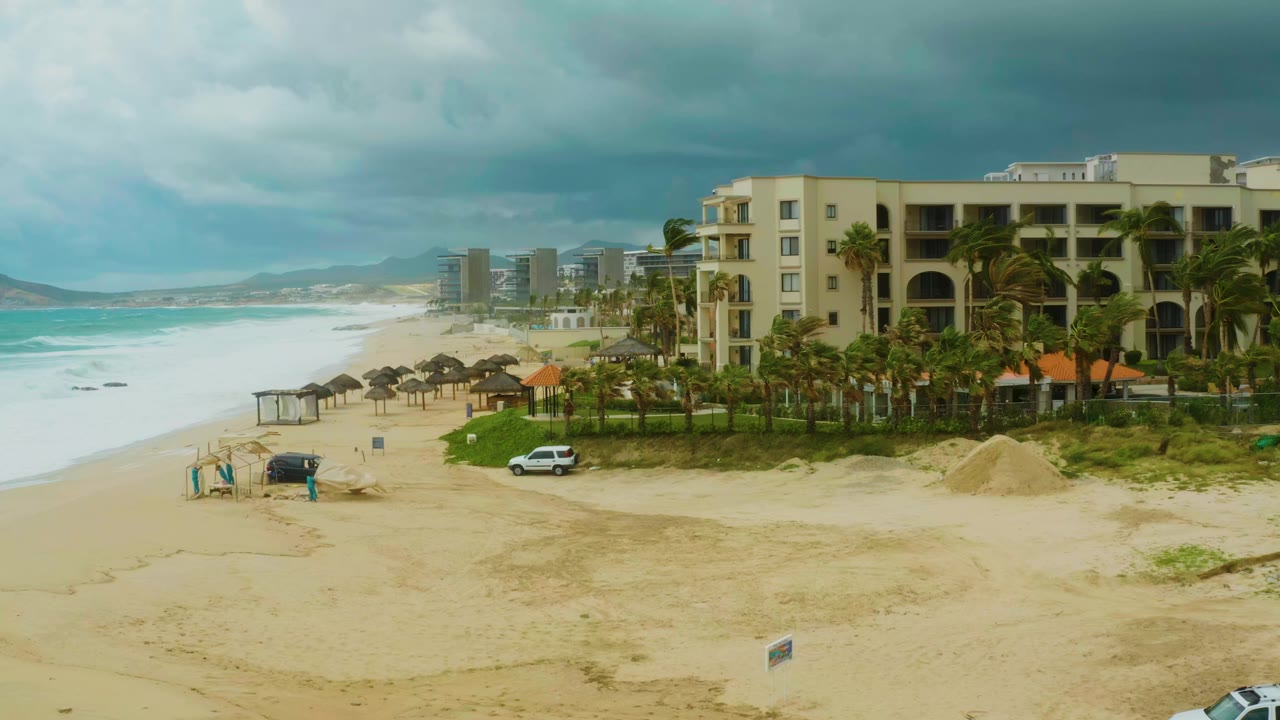 Elevated view of Dreams Resort Los Cabos with pale sand, turquoise waves, palm trees, shade umbrellas, a white parked car, and small people near a dark beach vehicle at 0:00.
