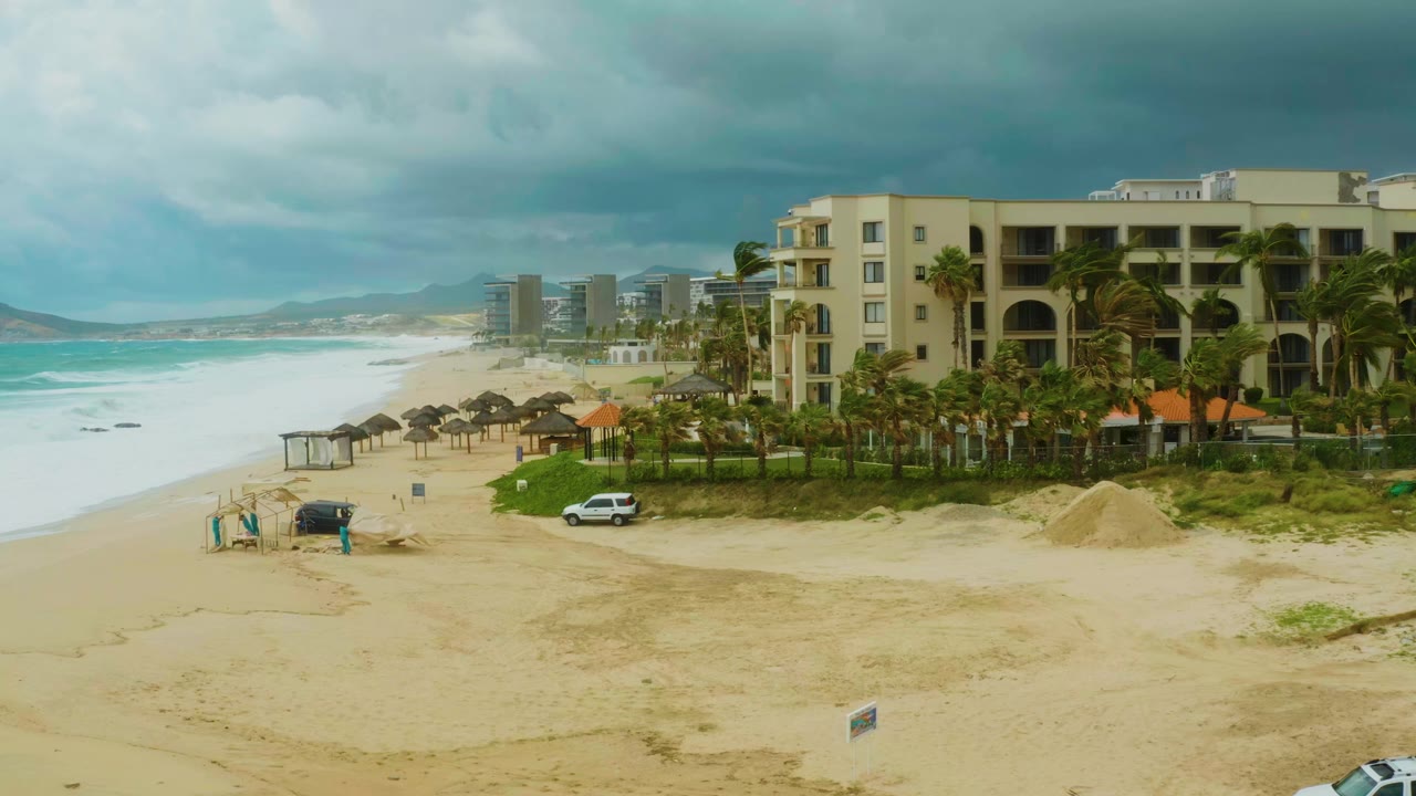 Dreams Resort Los Cabos beachfront remains in a wide elevated frame as waves roll along the left, the resort building fills the right, and the dark beach vehicle sits near small visible people.