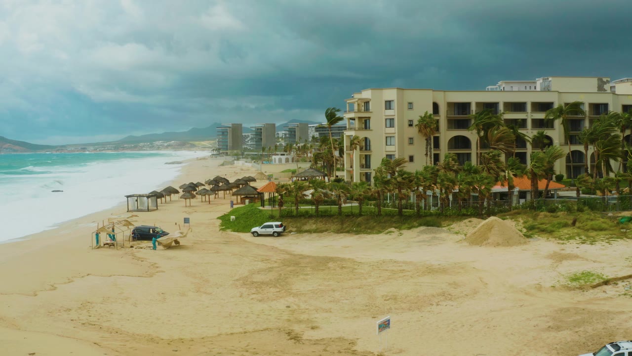 Elevated frame of Dreams Resort Los Cabos with surf breaking along the left edge, umbrella shade structures near the beach, a white car near palms, and small people by a dark vehicle.