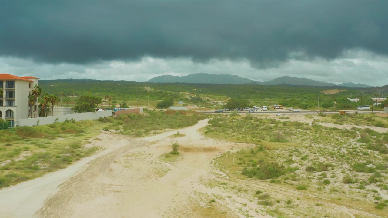 Dirt track through scrub with road and mountains beyond.