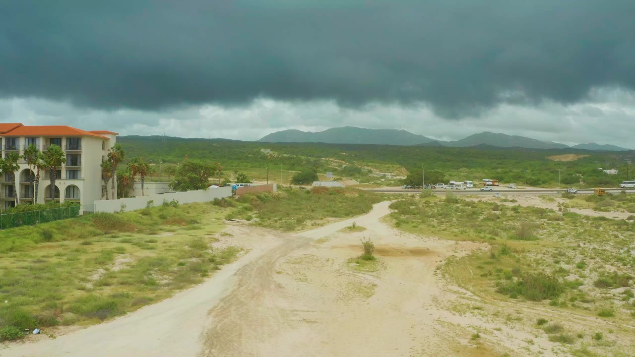 Opening frame at Dreams Resort Los Cabos showing a pale dirt road through scrubby ground, a tile-roofed building at left, distant highway traffic, low hills, and no visible people.