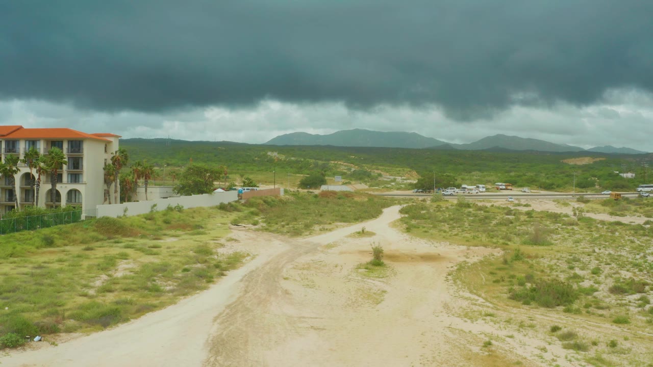 At 0:02, the elevated view at Dreams Resort Los Cabos shows the dirt track curving through open scrub, highway vehicles across the middle distance, hills beyond, and no visible people.