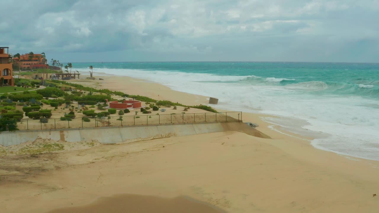 Opening frame of Dreams Resort Los Cabos beach with a broad sand foreground, turquoise waves breaking to the right, resort buildings at far left, and no visible people.