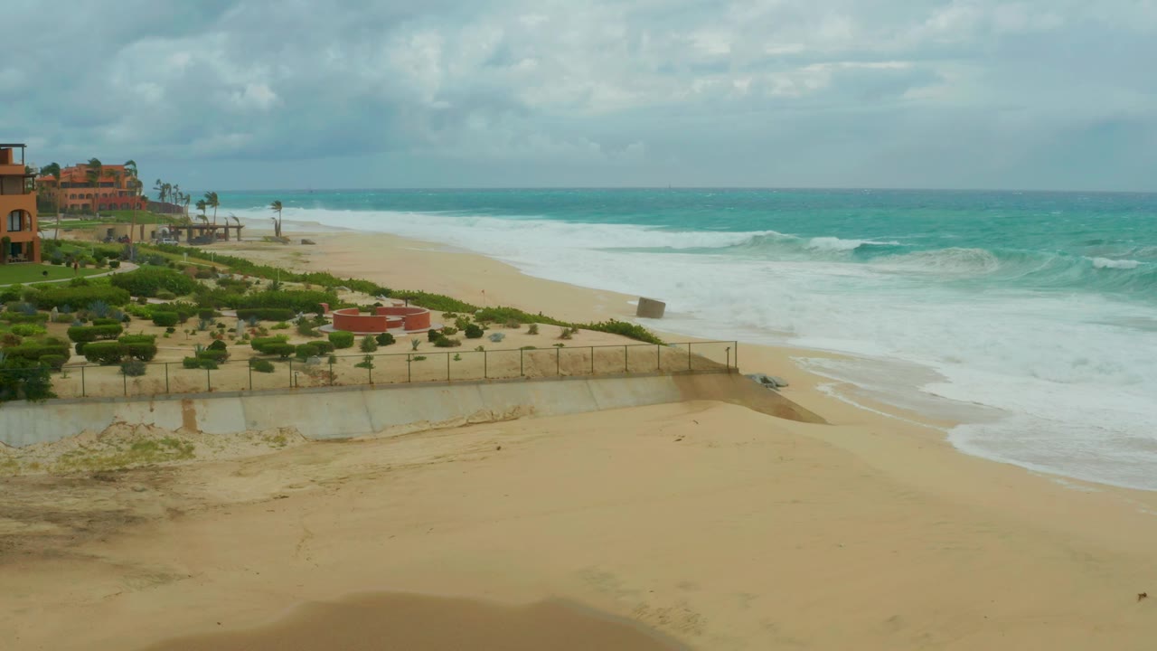 Dreams Resort Los Cabos shoreline with white foam pushing farther up the sand, the low wall and fence across the midground, and no visible people.