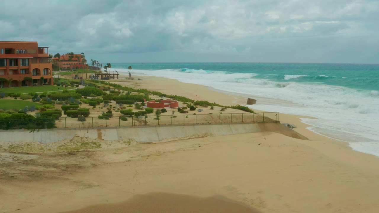 At one second, the Dreams Resort Los Cabos shoreline remains framed from above as a broad white wave band moves closer to the sand, with resort buildings and clipped greenery at left and no visible people.