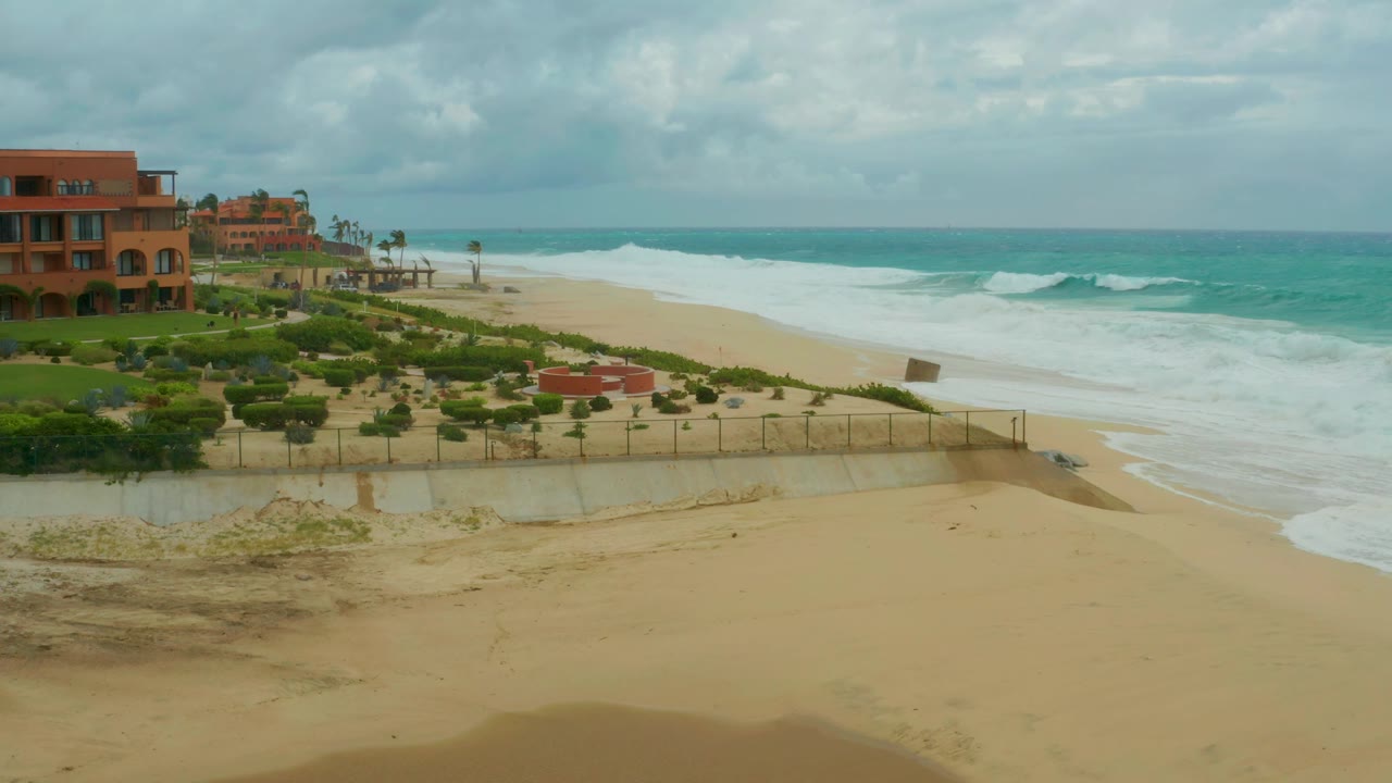 At three seconds, the elevated Dreams Resort Los Cabos view shows a darker cloud layer above turquoise water, a foamy surf break, empty sand, fencing, greenery, and coastal buildings at the left edge.