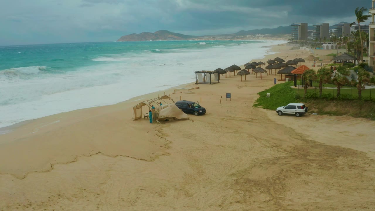 Elevated Dreams Resort Los Cabos shoreline view with surf pushing farther up the sand, parked black and white vehicles near shade structures, palms at right, and no visible people.