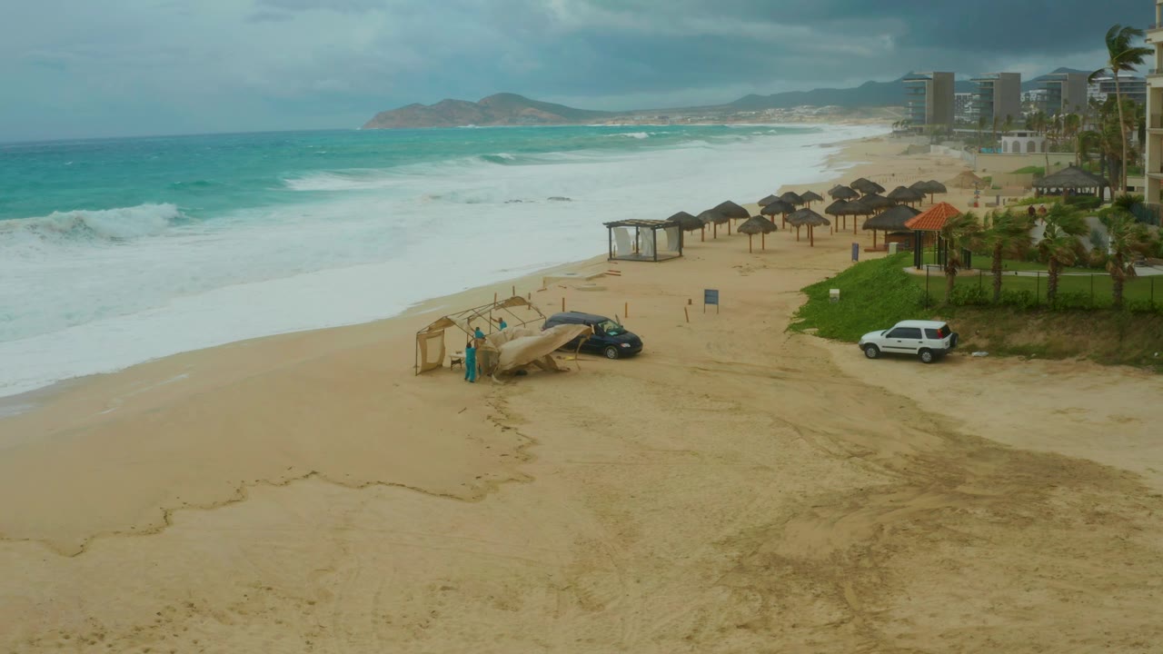 Dreams Resort Los Cabos beach frame with whitecaps across the left, a wide empty sand foreground, parked cars near a small beach structure, resort towers in the distance, and no visible people.