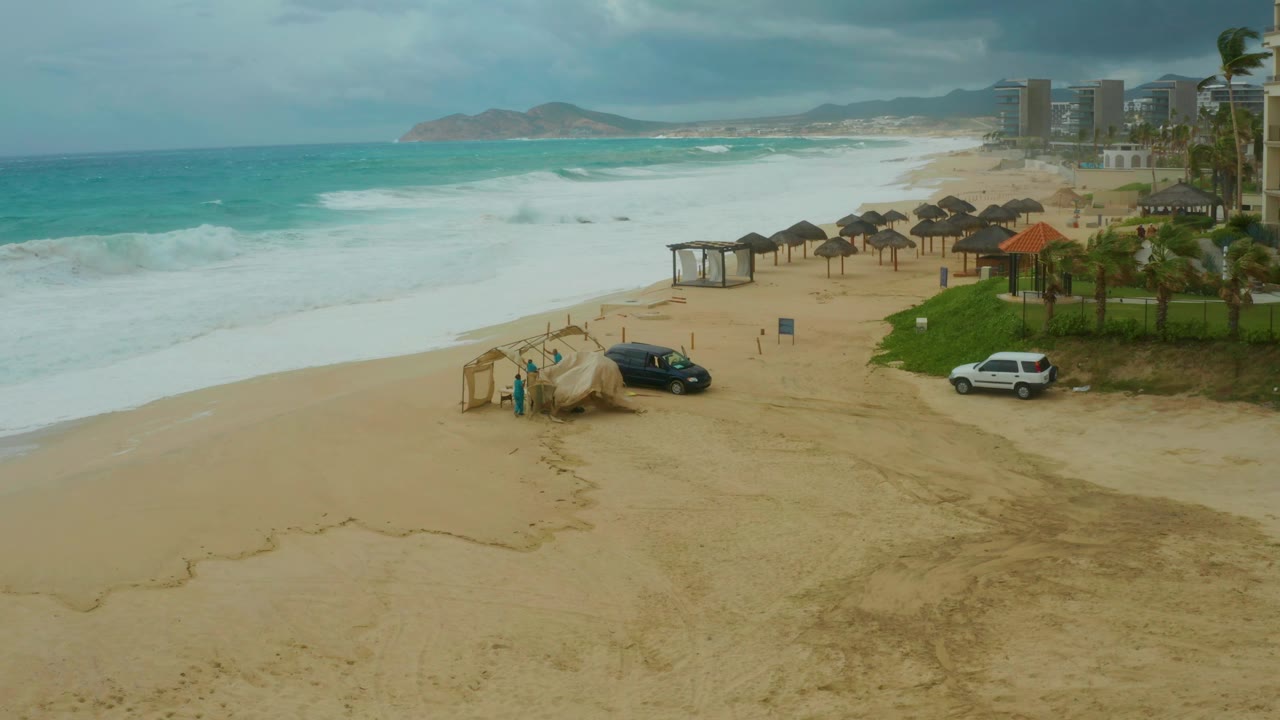 Elevated resort beach view in Los Cabos with a breaking wave near shore, thatched umbrellas arranged along the sand, two parked vehicles, palm trees, cloudy sky, and no visible people.