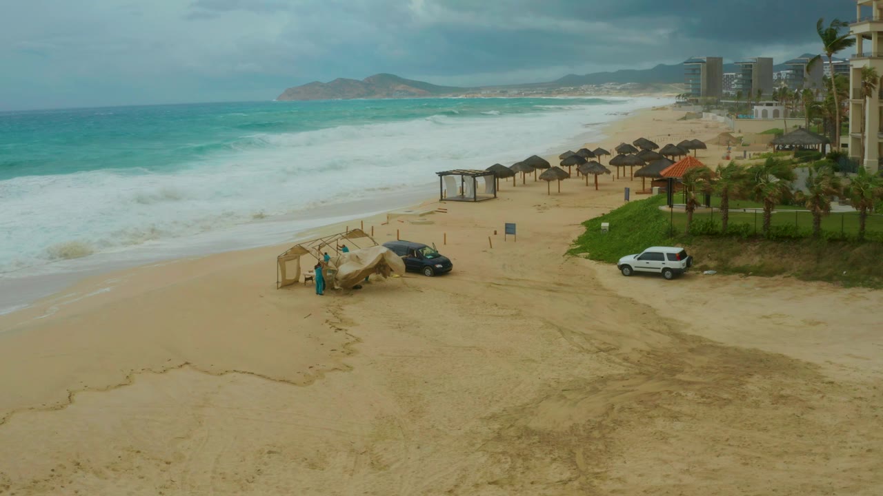 Dreams Resort Los Cabos shoreline under cloudy light, with foamy waves farther up the beach and two parked vehicles beside cabana structures, no visible people.