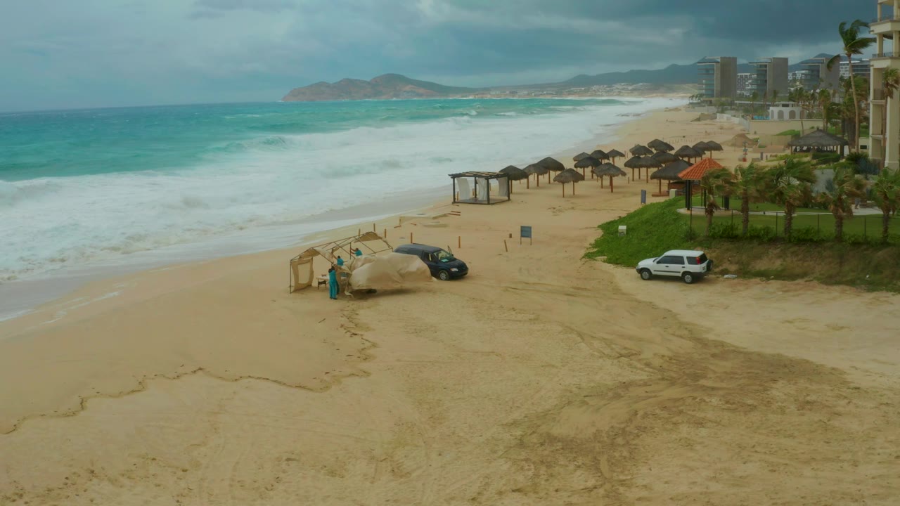 Elevated resort-beach view in Dreams Resort Los Cabos with rough surf, thatched cabanas, a dark car, a white SUV, palms, hotel buildings, and no visible people.
