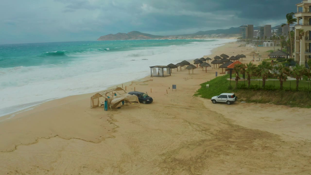 Opening view of Dreams Resort Los Cabos beach with turquoise waves offshore, thatched huts on the sand, parked vehicles near the foreground, resort buildings at right, and no visible people.