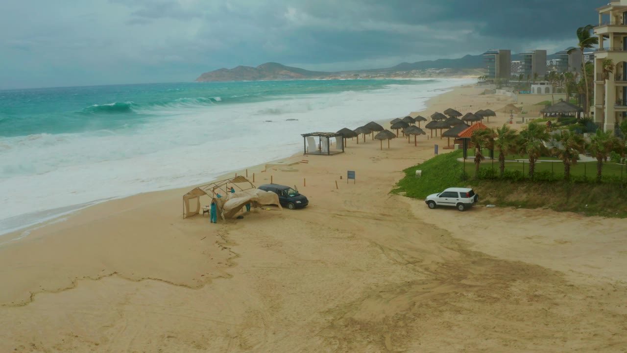 Beach surf breaking near thatched huts and parked vehicles under clouds.