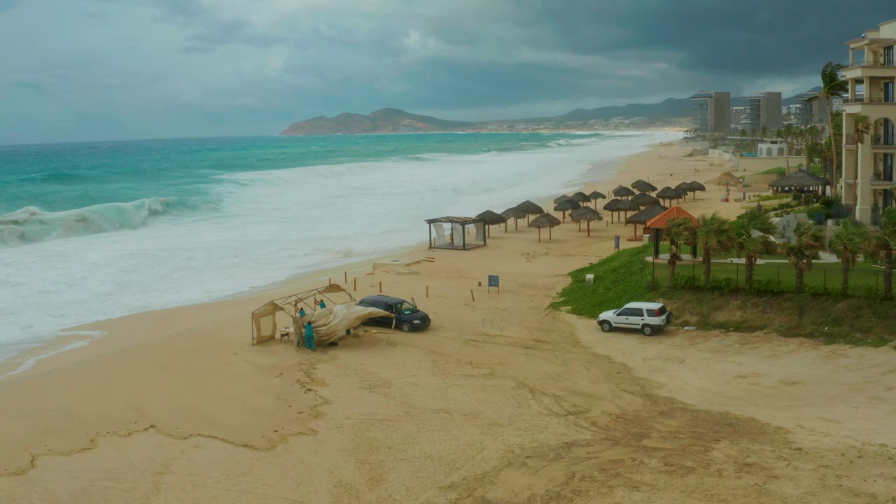 At Dreams Resort Los Cabos, white surf spreads onto the sandy beach below thatched umbrellas, two parked cars, beachfront buildings, distant hills, dark clouds, and small visible people near a beach structure.