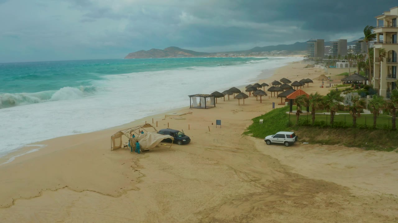 At 0:02, broad white foam covers more of the sand at Dreams Resort Los Cabos, with thatched umbrella rows, a beach structure, parked cars, resort buildings, distant hills, and small visible people near the foreground structure.