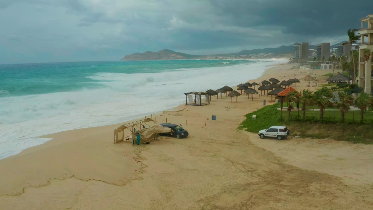 Opening view of Dreams Resort Los Cabos beach with white surf spreading across the sand, palapa umbrellas and buildings to the right, parked vehicles near the beach access, and no visible people.
