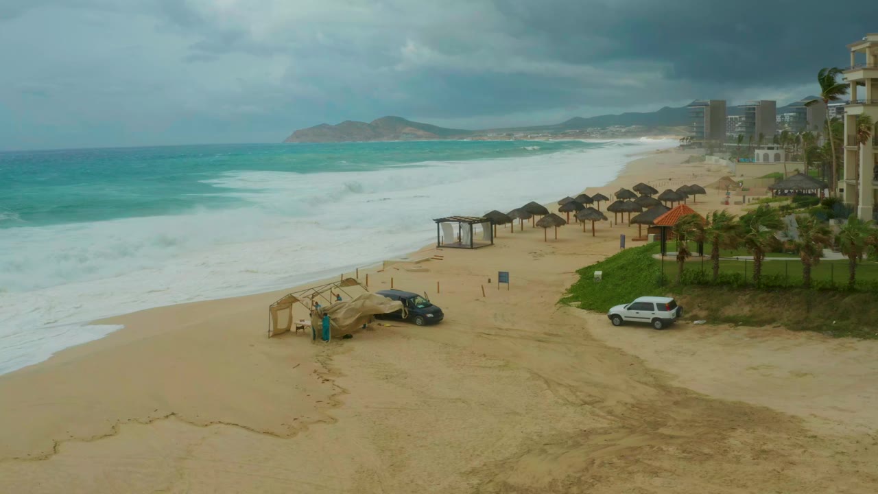 At 0:01, Dreams Resort Los Cabos beach remains framed from above as the surf line advances, with palapa umbrellas, palms, resort buildings, two parked vehicles, and no visible people.