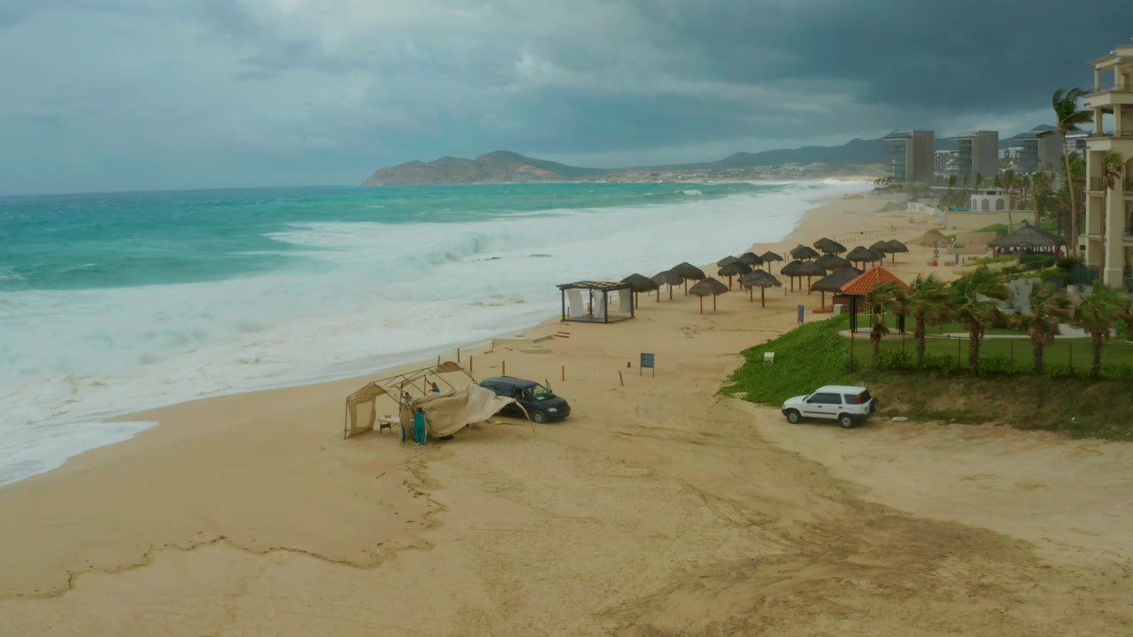 At 0:02, the Dreams Resort Los Cabos shoreline shows a broad sheet of white water in the midground, darker clouds overhead, resort towers on the right, parked cars below, and no visible people.