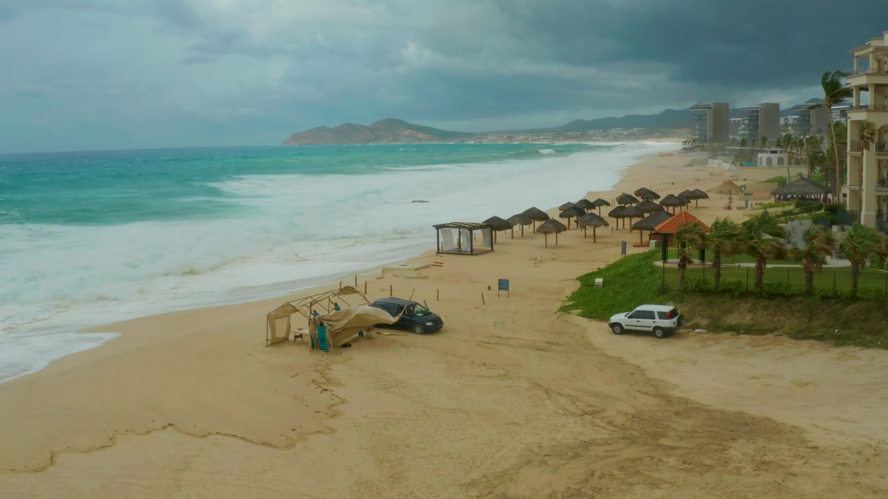 Beachfront resort scene with large surf, umbrellas, palm trees, parked vehicles, and cloudy sky.