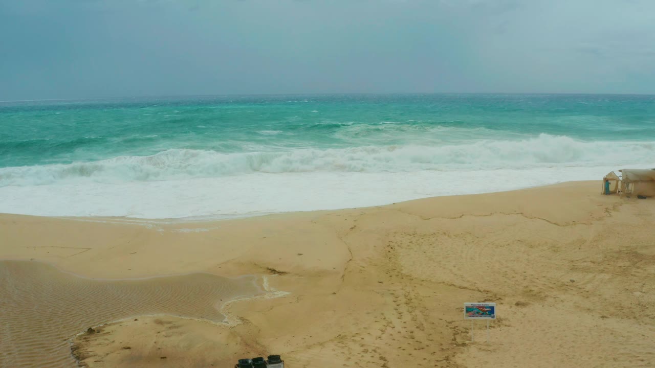 Opening frame at Dreams Resort Los Cabos shows turquoise surf breaking onto a broad sandy beach with a small blue sign low in frame and no visible people.
