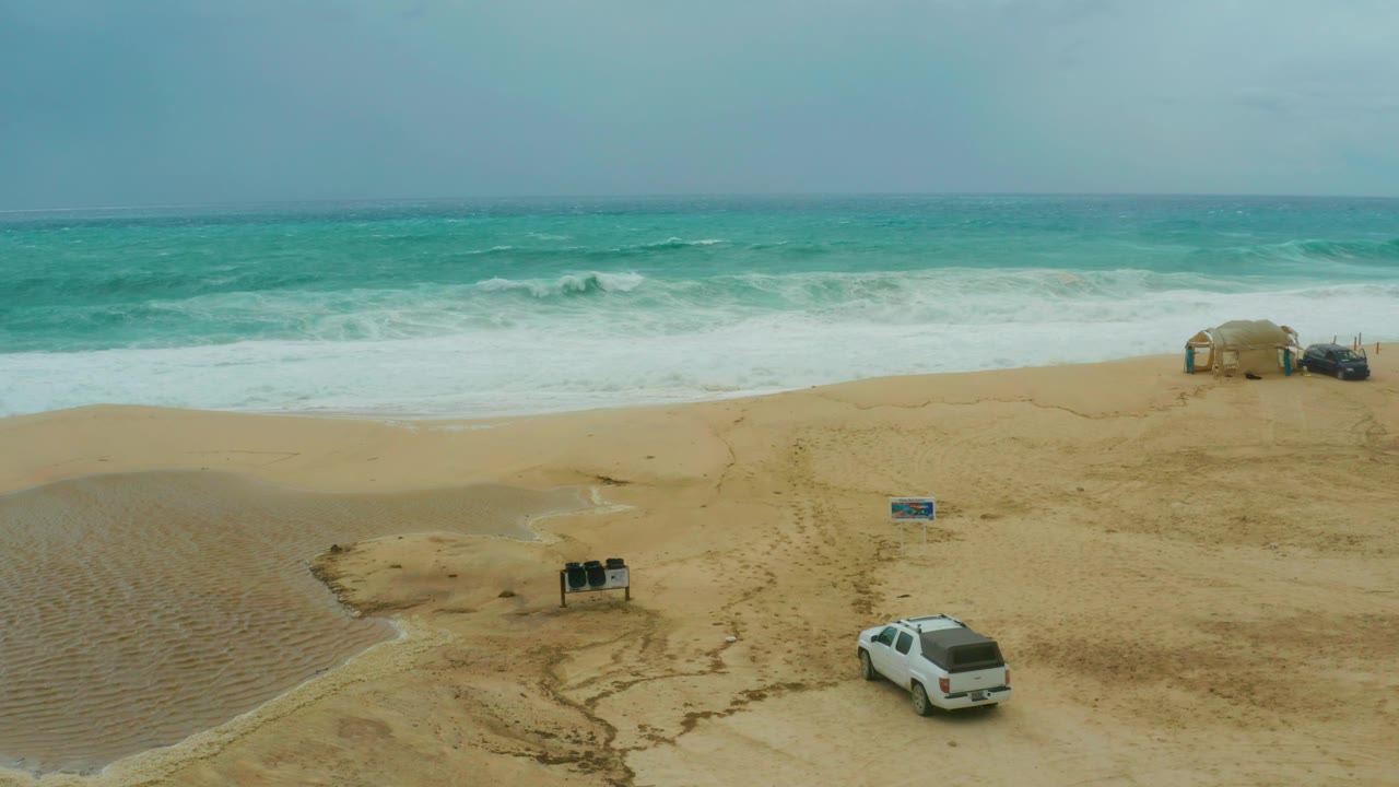 The white SUV has shifted farther right on the sandy beach while small signs remain near the center and white surf rolls across the background, with no visible people.