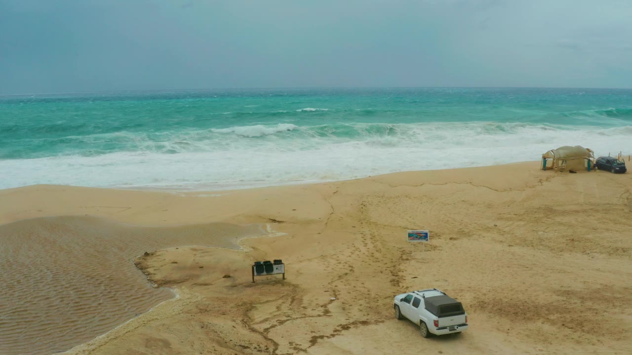 The SUV nears the lower right edge of the aerial frame as waves wash toward the open tan beach and the shade structure remains near the upper right, with no visible people.