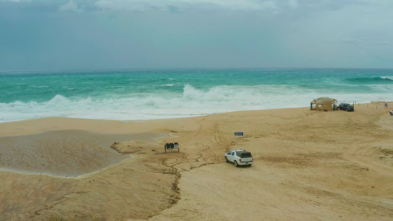 At Dreams Resort Los Cabos in Baja California Sur, a white SUV sits low in frame on pale sand while surf breaks across the background and no visible people appear.