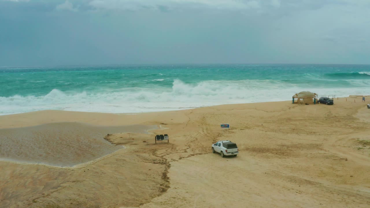 The white SUV has advanced slightly across the sandy beach at Dreams Resort Los Cabos, with turquoise surf rolling behind it and no visible people in the frame.