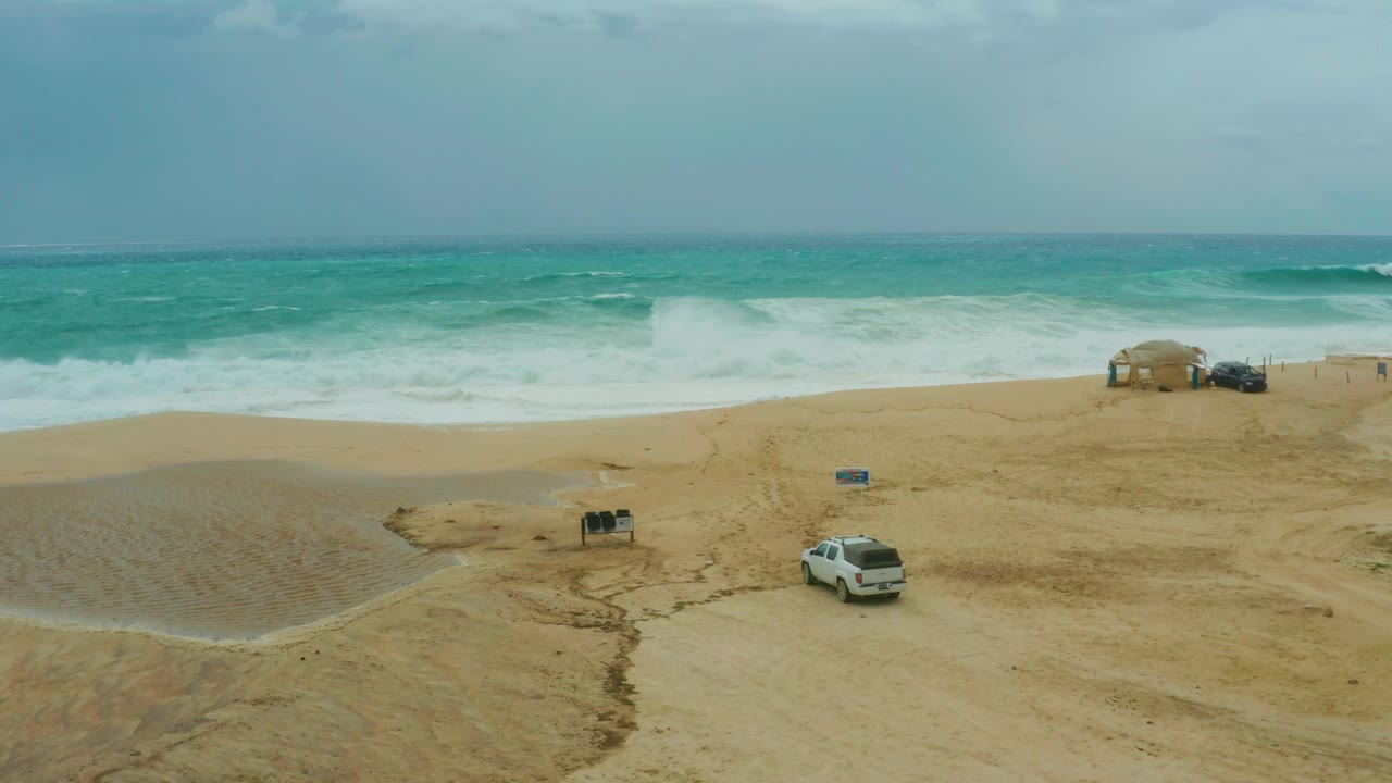 An elevated view at Dreams Resort Los Cabos shows the white SUV farther along the beach, with a wet sand channel at left, surf across the top, and no visible people.