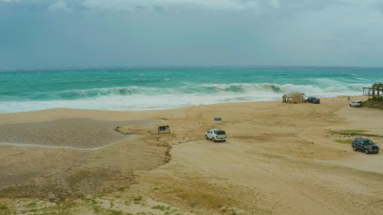 Wide elevated view of Dreams Resort Los Cabos beach with turquoise waves breaking behind parked vehicles on sand, no visible people.