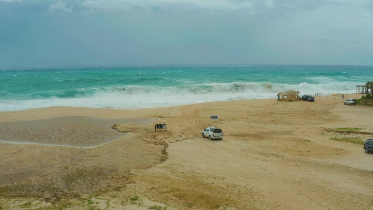 Elevated frame of Dreams Resort Los Cabos beach where rough surf spreads along the shore and vehicles sit near sand tracks, no visible people.