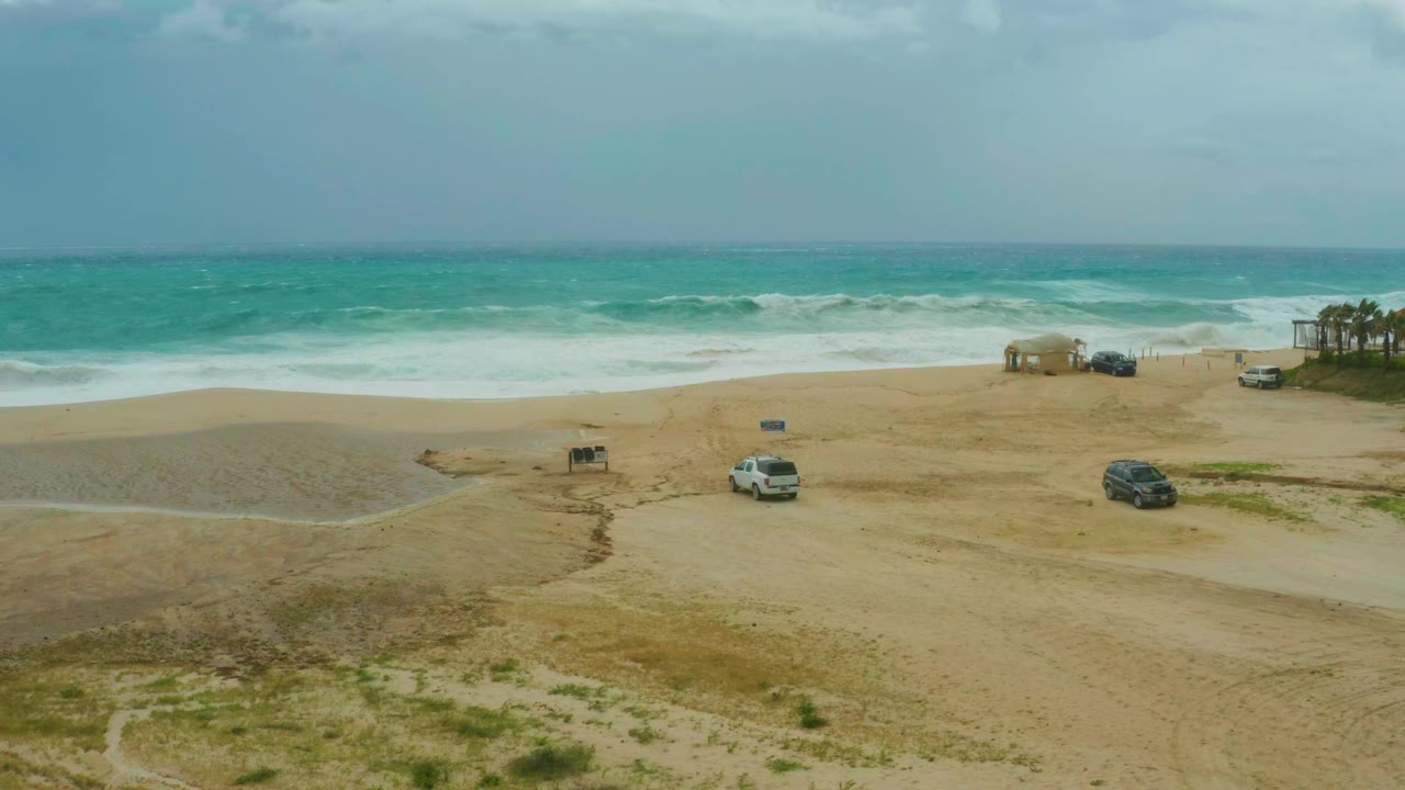 Aerial frame over Dreams Resort Los Cabos beach with white surf rolling toward the sand, vehicles spaced across the midground, and no visible people.