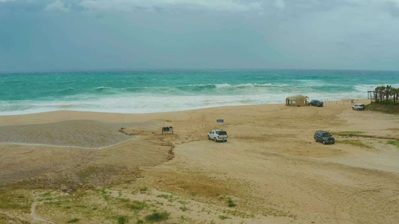 High wide view of the Dreams Resort Los Cabos shoreline, with overcast sky above blue-green water, a white vehicle on the sand, a dark vehicle farther right, and no visible people.