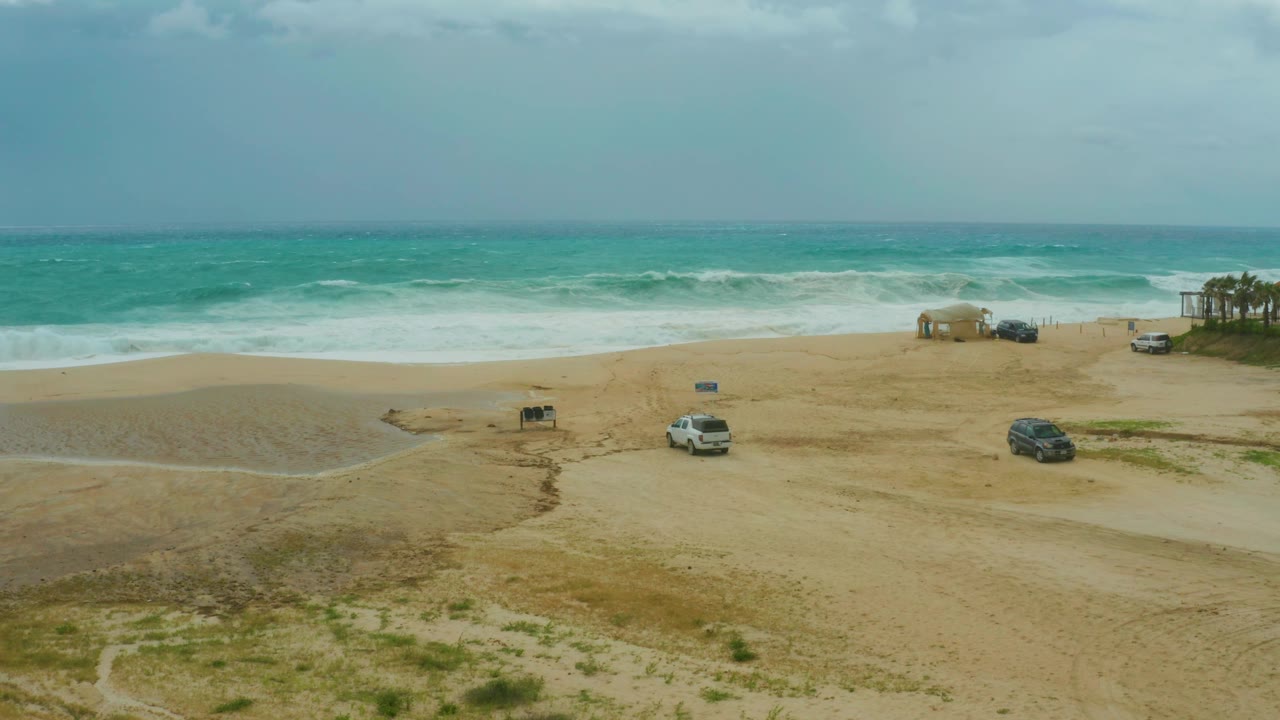 Mid-clip aerial view at Dreams Resort Los Cabos, Mexico, showing rolling surf, pale sand, a white vehicle near the lower center, a dark vehicle to the right, and no visible people.