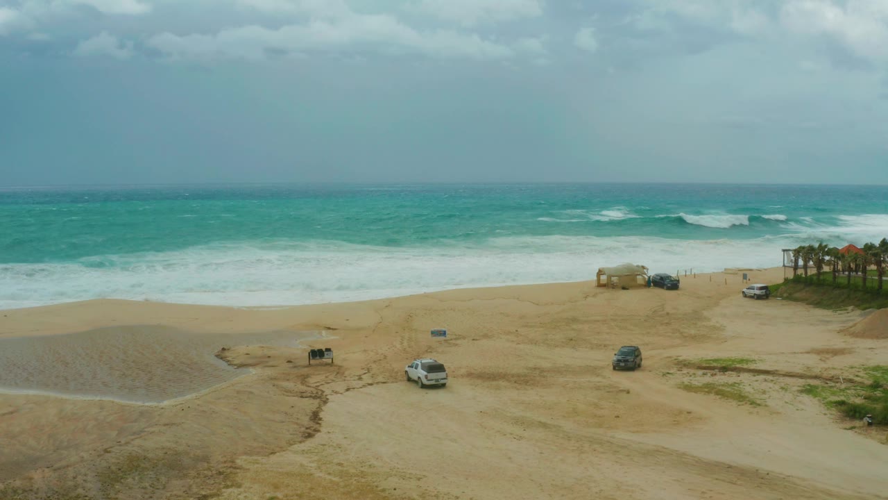 Dreams Resort Los Cabos shoreline with the white SUV near the lower sand, darker vehicles closer to the beach access area, rolling surf, and no visible people.