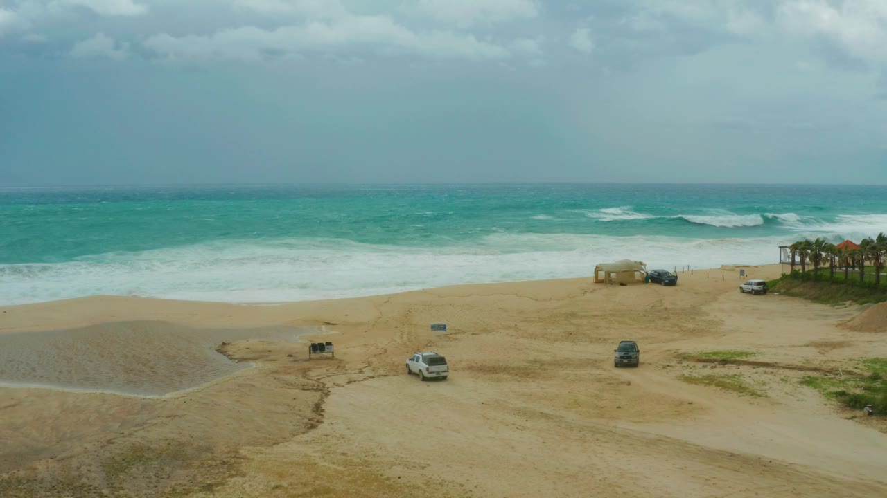Wide view at Dreams Resort Los Cabos showing the same sandy beach, a white SUV left of center, a dark vehicle to the right, choppy ocean waves, and no visible people.