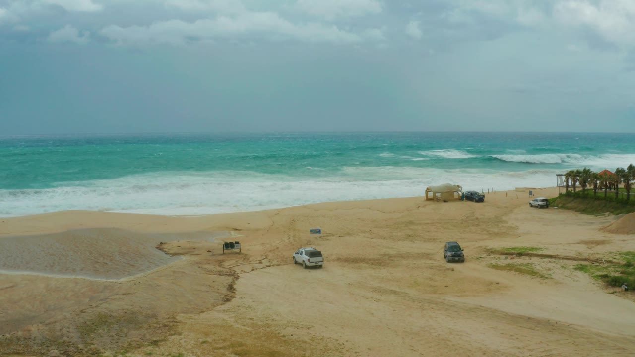 At 0:03, Dreams Resort Los Cabos beach remains empty of visible people, with turquoise water, white surf, parked vehicles, and a dark vehicle slightly farther along the sand.