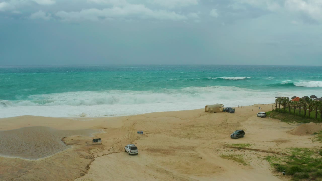 Overcast beach with turquoise waves, parked vehicles, sand dunes, and palms.