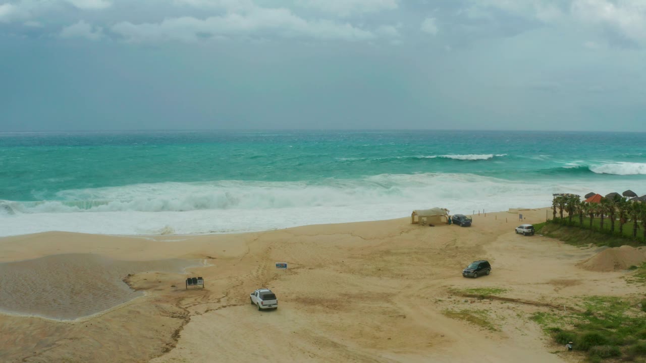 The Los Cabos beach remains empty of visible people as white surf spreads across the shore and parked vehicles sit near a sandy access area.