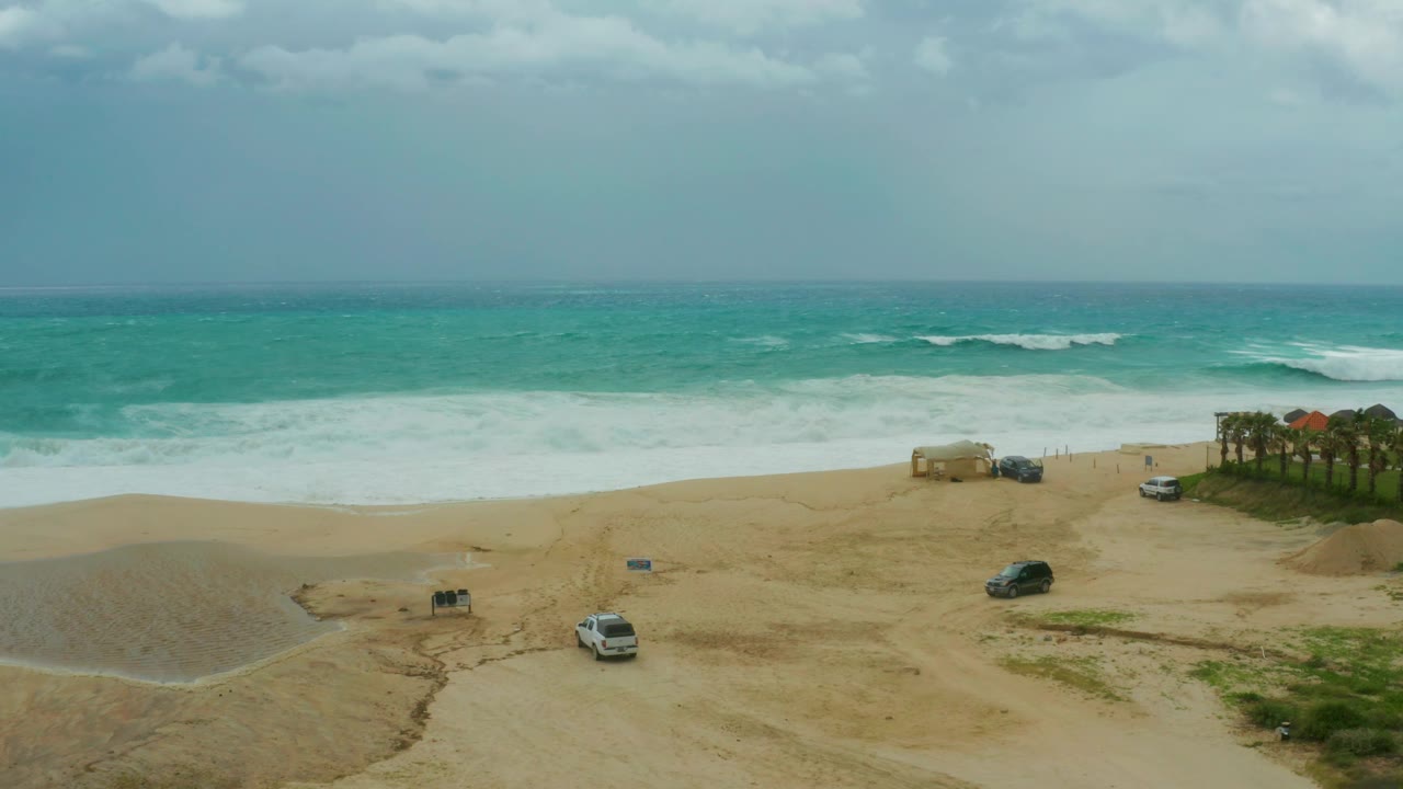 The elevated view shows rough turquoise surf meeting pale sand, with vehicles spaced across the beach area and no visible people at Dreams Resort Los Cabos.