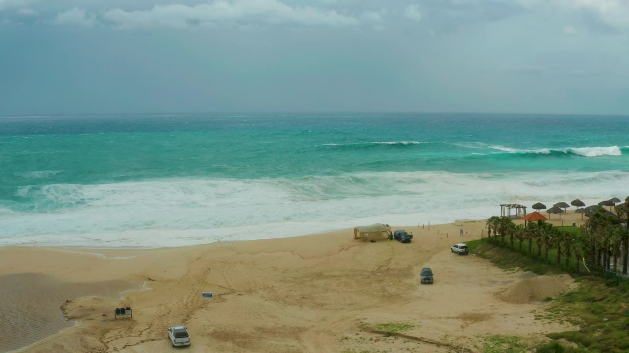 Wide aerial beach view with turquoise surf, sandy access area, vehicles, and palm trees.