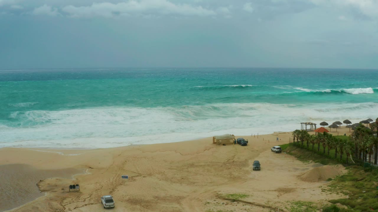 At 0:01, the Dreams Resort Los Cabos shoreline holds a broad band of white surf while a white van and darker vehicles sit or move slowly across the sand, with no visible people.