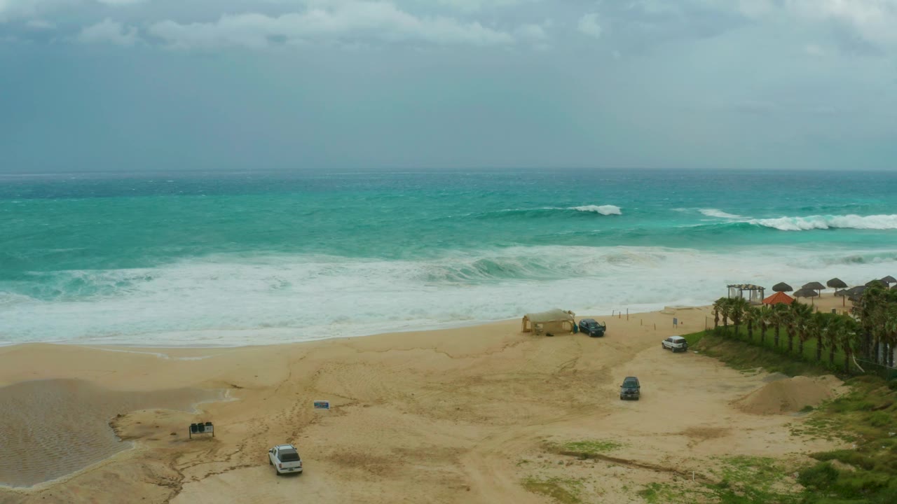 At 0:03, a darker turquoise ocean and white surf sit behind a sandy foreground with a white van, a dark vehicle near center, palm trees at right, and no visible people.