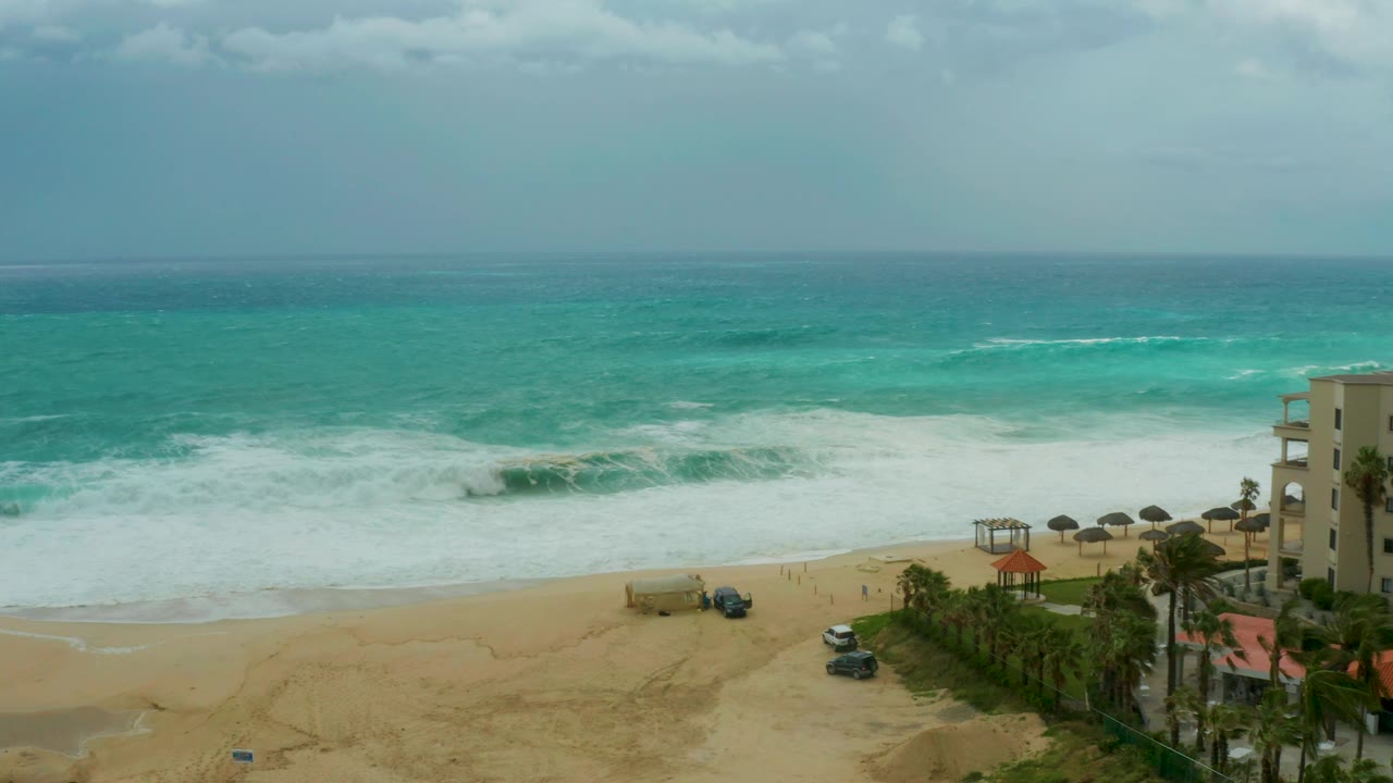 At 0:01, Dreams Resort Los Cabos beach remains framed from above as a larger wave line approaches the sand, with vehicles below, umbrellas and palms at right, and no visible people.