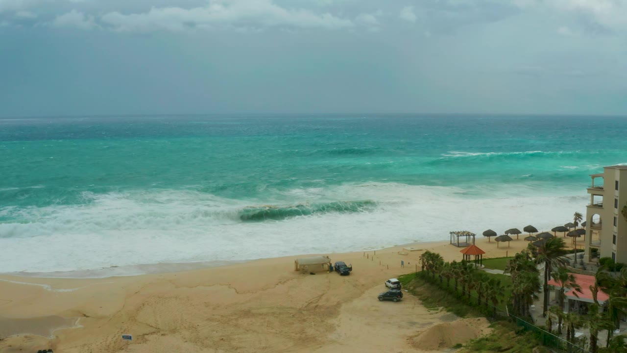 At 0:02, the surf spreads wider across Dreams Resort Los Cabos beach, with a white car and darker vehicle on the sand, palm-lined resort frontage at right, and no visible people.