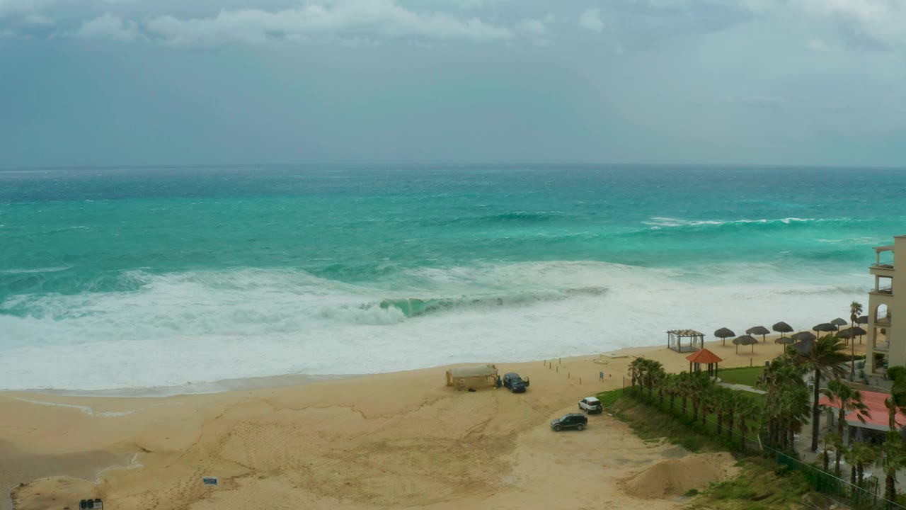 Aerial view of cloudy ocean surf over a sandy beach with cars and palm-lined resort frontage.
