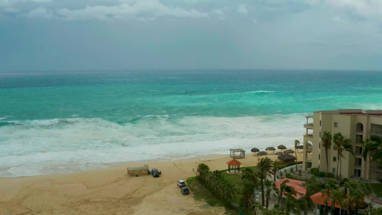 At 0:01, white foam spreads wider across the Dreams Los Cabos shoreline while vehicles remain on the sand near palms and the right-edge resort building, with no visible people.