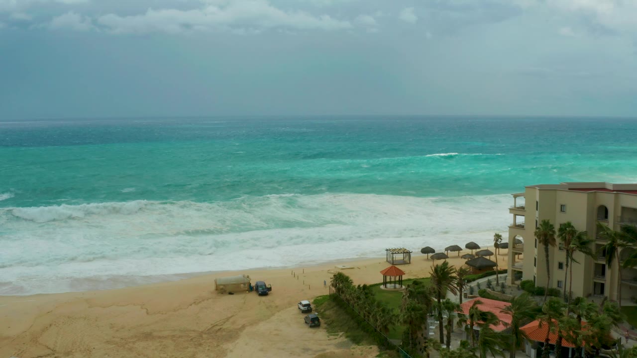 At 0:03, broad white foam covers more of the nearshore water at Dreams Los Cabos, with vehicles on the beach access area and palms framing the resort building, with no visible people.