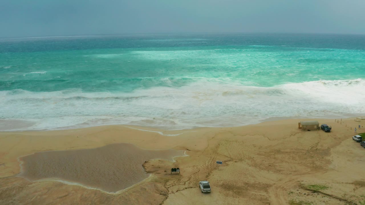 At Dreams Resort Los Cabos, turquoise water and white surf meet tan sand while a tidal pool, parked vehicles, and a small beach structure are visible with no visible people.